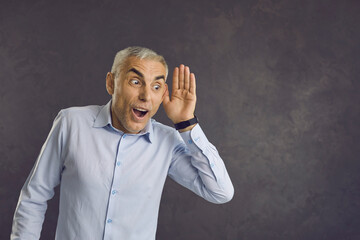 Older man eavesdrops on secret information or listens to hot news and shocking gossip. Man standing on gray background with funny expression puts his hand to his ear listening. Banner. Place for text.