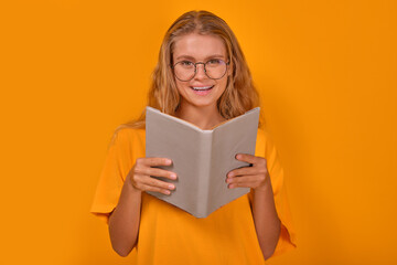 Young cheerful laughing Caucasian woman bookworm with glasses reads book from library or bookstore and giggles enjoying funny stories from mood-improving literature stands on yellow background.