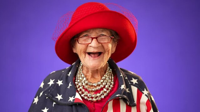 Closeup portrait of smiling, happy, funny crazy grandmother mature woman, 80s, wearing patriotic flag jacket isolated on purple background. Concept of youthful old female.