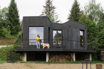 Woman wearing yellow coat and blue jeans and golden retriever dog near modern wooden cabin.