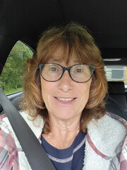 Mature woman taking a selfie while sitting in a parked car. She is smiling at the camera and wearing her seat belt.