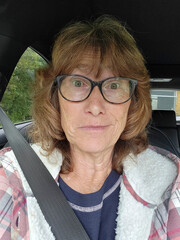 Mature woman taking a selfie while sitting in a parked car. She is making a funny face and is still wearing her seat belt.
