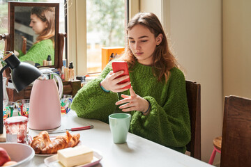 Lovely serene teen woman using her smartphone while relaxing before breakfast - obrazy, fototapety, plakaty
