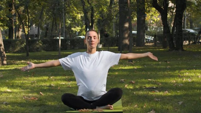 Young pregnant woman sitting on a yoga mat in a lotus pose and slowly raising her hands to make a namaste gesture close up on a sunny day in a city park. Concept of benefits of meditation for a