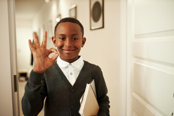 Brilliant successful african american school kid with copybook in hands showing ok sign with fingers standing in doorway, sharing good news with parents after passing exam and getting excellent mark