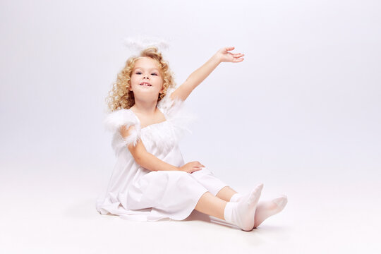 Beautiful little girl, child with curly hair in image of cute angel sitting on floor isolated over white studio background. Concept of childhood, imagination, fantasy, fashion and beauty, holidays