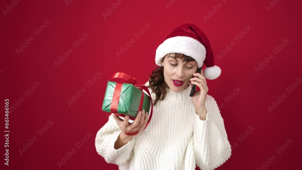 Sticker young caucasian woman smiling wearing christmas hat holding gift speaking on the phone over isolated