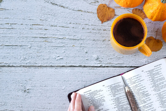Hand holding open holy bible book of psalms with coffee cup, autumn leaves, and pumpkin on wooden table. Top view. copy space. Christian thanksgiving, biblical study, gratitude and praise concept.