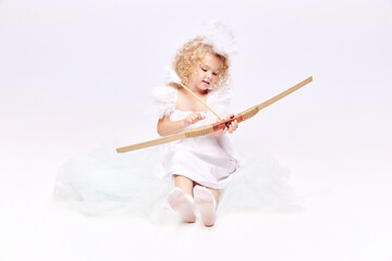 Fototapeta premium Little baby cupid, beautiful girl in image of angel holding bow isolated over white studio background. Valentine's day. Concept of childhood, imagination, fantasy, fashion and beauty, holidays