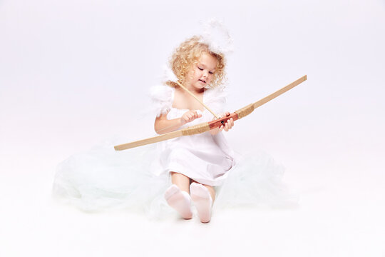 Little baby cupid, beautiful girl in image of angel holding bow isolated over white studio background. Valentine's day. Concept of childhood, imagination, fantasy, fashion and beauty, holidays