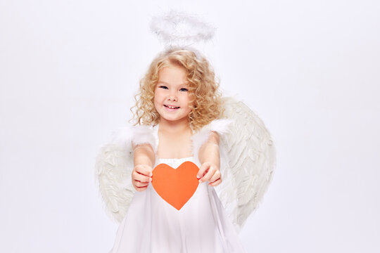 Happy Valentine's Day. Little baby cupid, beautiful girl in white dress holding paper heart isolated over white studio background. Concept of childhood, imagination, fantasy, happiness, holidays