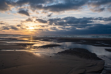 A breathtaking moment captured as the golden sun sets over a picturesque French beach, casting warm hues on the shoreline, with small waves gently rolling in under dramatic clouds. Golden Sunset at
