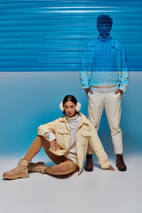 asian woman in warm earmuffs sitting near man with hands in pockets and blue plastic sheet in studio - obrazy, fototapety, plakaty