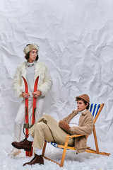 man in deck chair near trendy asian woman in skis on snow in studio, winter fashion and leisure