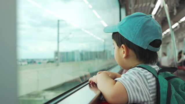 Little Asian Japanese boy in backpack and blue cap looking through the window while going to the school by commuter train.