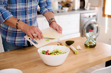 Close-up hands of chef putting chopped avocado fruit into a bowl of healthy salad while preparing dinner in the kitchen