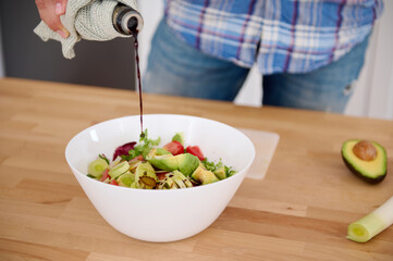 Detail on the hands of chef pouring balsamic vinegar into a bowl of fresh salad while cooking in minimalist home kitchen