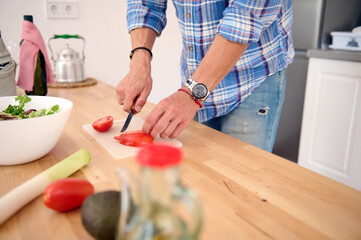 Close-up of male hands cutting fresh ripe organic juicy tomatoes while preparing salad in home kitchen. Healthy eating