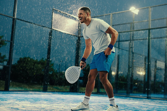 Happy padel athlete standing on the court with his racket on a rainy evening