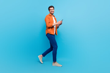Full body profile portrait of attractive cheerful person use smart phone walking empty space isolated on blue color background