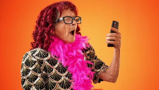 Funny crazy happy elderly old toothless woman taking selfie using mobile cell phone wearing boa and glasses isolated on solid orange background studio portrait. 