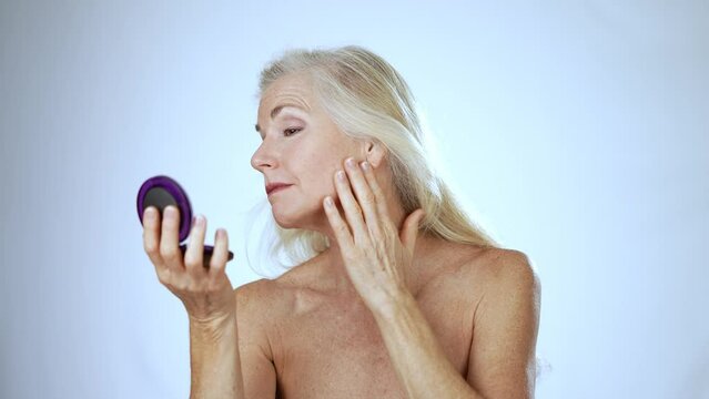 Mature middle aged nude woman looking at her blonde gray hair in compact mirror considering face lift and smiling isolated on white background with copy space.