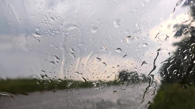 Raining water on glass windscreen window, drops close-up on road sidewalk with cloudy blurred background. Moody rainy footage