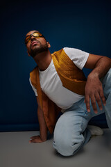 stylish african american man in vest and sunglasses looking up on blue backdrop, fashion concept