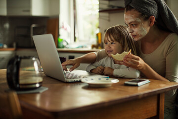 Young mother wearing a facial mask while using a laptop with her daughter at home