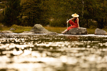 Woman with straw hat in middle of forest with river in background trees and nature warm colors, meditating in nature, traveling through east