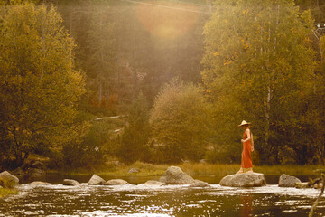 Fototapeta premium Woman with straw hat in middle of forest with river in background trees and nature warm colors, meditating in nature, traveling through east