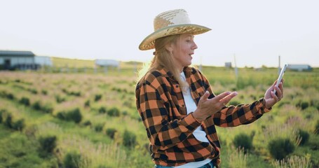 Middle-aged beautiful woman farmer in a hat has online conference with agribusiness partners standing in a field at sunset. Agricultural specialist video call by phone.