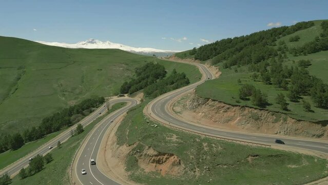 Steep serpentine between hills in Lori in Armenia. Cars are driving along the highway. There is grass and trees growing on the mountains. Behind the snowy mountain Aragats. Drone video. Fly forward.