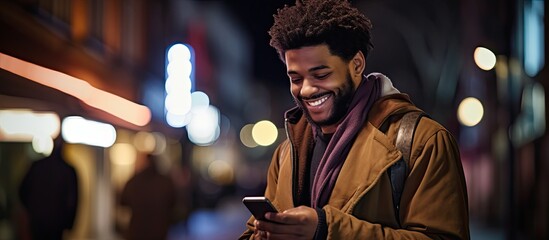 An African American man is using his phone on a city street at night while looking away - obrazy, fototapety, plakaty