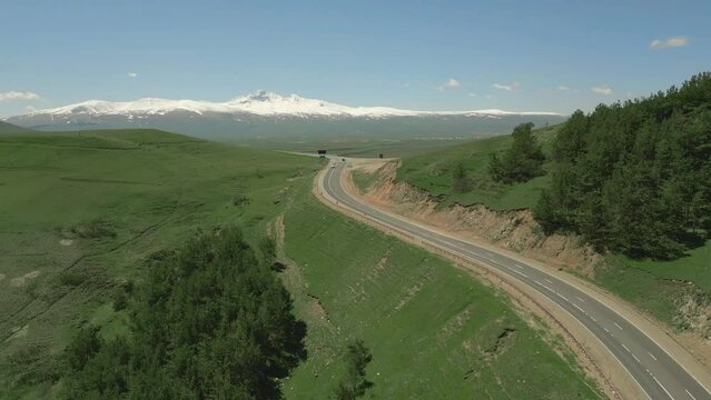 Steep serpentine between hills in Lori in Armenia. Cars are driving along the highway. There is grass and trees growing on the mountains. Behind the snowy mountain Aragats. Drone video. Fly forward.