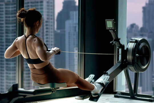 Fitness girl workout on indoor rower at the gym, doing exercises on rowing machine near the window,  view of skyscrapers in the background, woman exercising in fitness club