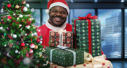 African Santa holds gift boxes behind a Christmas tree near the window, blurred high buildings in ...