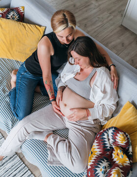 Young woman tender touching partner's female pregnant belly. Same-sex marriage couple in the home living room. Woman's health, happy pregnancy doula supporting and calm mental mood concept image.