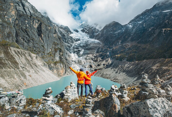 Couple trekkers dressed bright jackets on the rock enjoying a glacier falling in high altitude Sabai Tso glacial lake cca 4350m. Makalu Barun National Park, Mera peak climbing route, Nepal.