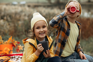 Little cute smiling girl and boy sitting near by car on plaid and drinking cocoa. Kid resting with her family in the nature. Picnic. Autumn season.