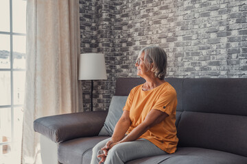 Obraz premium Positive thoughtful pretty middle aged blond woman sitting on home sofa, keeping hands at chin, looking at window away with good thoughts, thinking over future retirement, planning, dreaming.