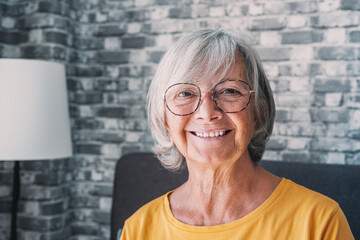 Smiling middle aged mature grey haired woman looking at camera, happy old lady in glasses posing at home indoor, positive single senior retired female sitting on sofa in living room headshot portrait. - obrazy, fototapety, plakaty