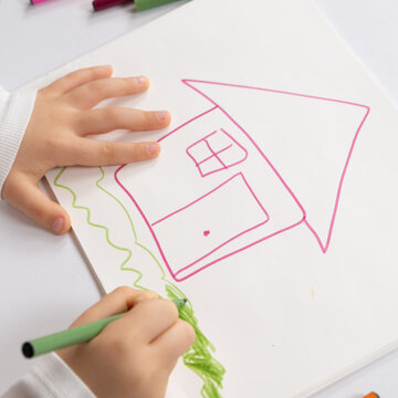 Little child drawing with felt-tip pens a house with grass in a sketchbook.