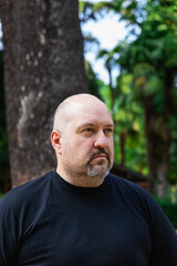 Fototapeta premium Portrait of a bald mature man with a goatee very serious in a park with a blank stare. Outdoor vertical portrait in natural light.
