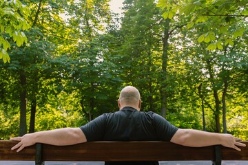 View from behind of mature man with little hair or alopecia sitting on a park bench.  Outdoor...
