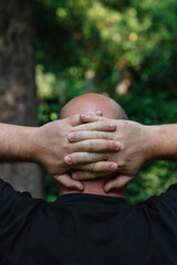 Fototapeta premium View from behind of mature man with little hair or alopecia with his hands on the nape of his neck. Outdoor vertical photography outdoors with natural light. Concept of reflection and observation 