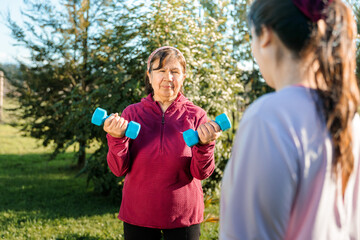 Latin senior woman doing exercise with dumbbells and being helped by a trainer outdoors on the grass.