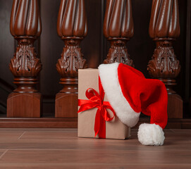 Obraz na płótnie Canvas Gift in a craft box with a red ribbon and Santa's hat in the house on the background of a wooden staircase