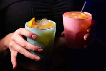 Beautiful girl in a nightclub in a black blouse holds in her hands two glasses with green and pink cocktails