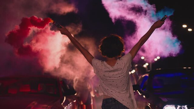 Young woman standing in middle of sport cars and waving , holding colorful smoke bombs flares . Girl starting muscle car drag racing at night . Back view slow motion . Ready for racing . Crowd of fans